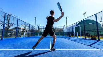 Low-angle view of a player in black swinging his padel racket on a bright blue outdoor court under a clear sky