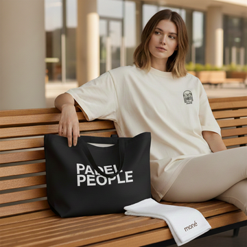 Woman sitting on a bench holding a black tote bag with 'PADEL PEOPLE' text, wearing a white t-shirt with a logo. Mone Padel | Moné 950ml Water Bottle | “Padel People” Tote Bag