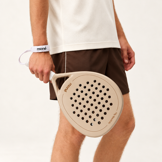 A man in a white T-shirt and brown shorts holds the beige Sahara Hybrid padel racket at his side, strap on wrist