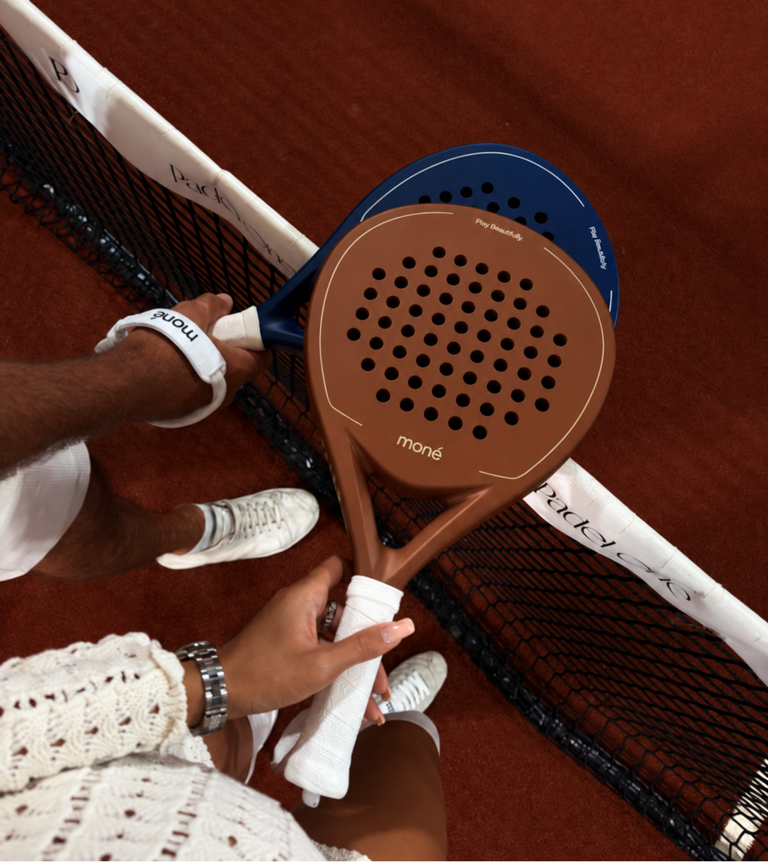 Two players holding brown and navy blue moné padel rackets together above the net on a red court.