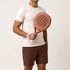 A man in a cream T-shirt and brown shorts holds the Sienna Hybrid padel racket in light brown against a light backdrop
