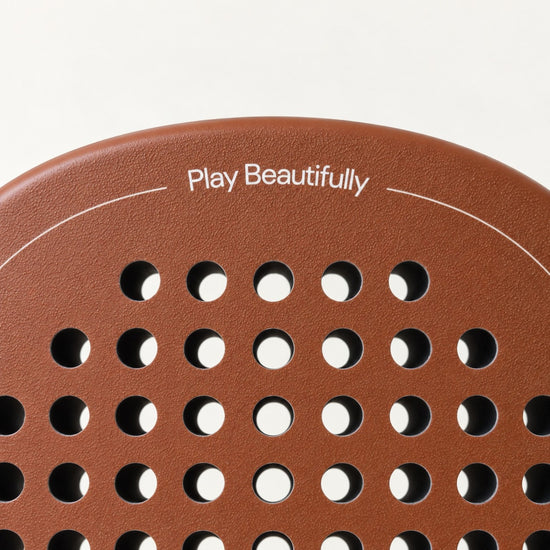 Play Beautifully printed in cream at the top of the light brown Sienna Hybrid padel racket, above its hole pattern